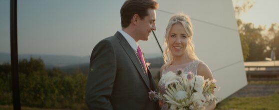 Martina und Lukas stehen bei Sonnenuntergang vor der modernen Fassade am Hannersberg, im Hintergrund die Weinberge – Szene aus ihrem Hochzeitsfilm. Hannersberg hochzeit videograf